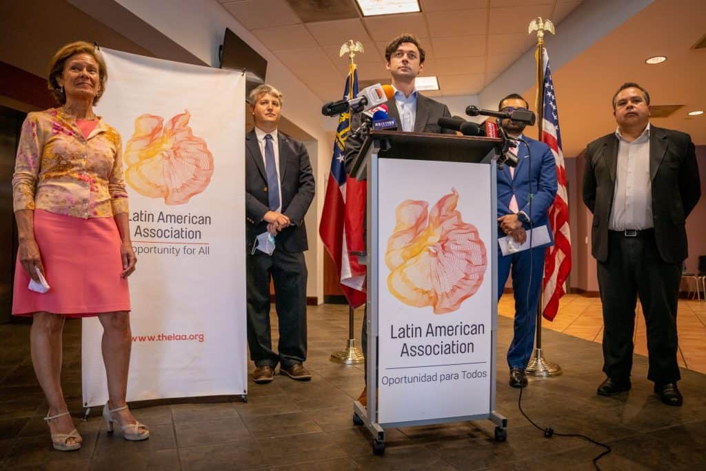 Senator Ossoff speaking at a podium at the Latin American Association in Atlanta with members of the organization beside him.