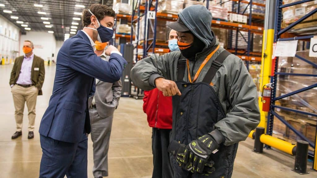 Senator Ossoff bumping elbows with a worker in a large warehouse with other staff members in the background.