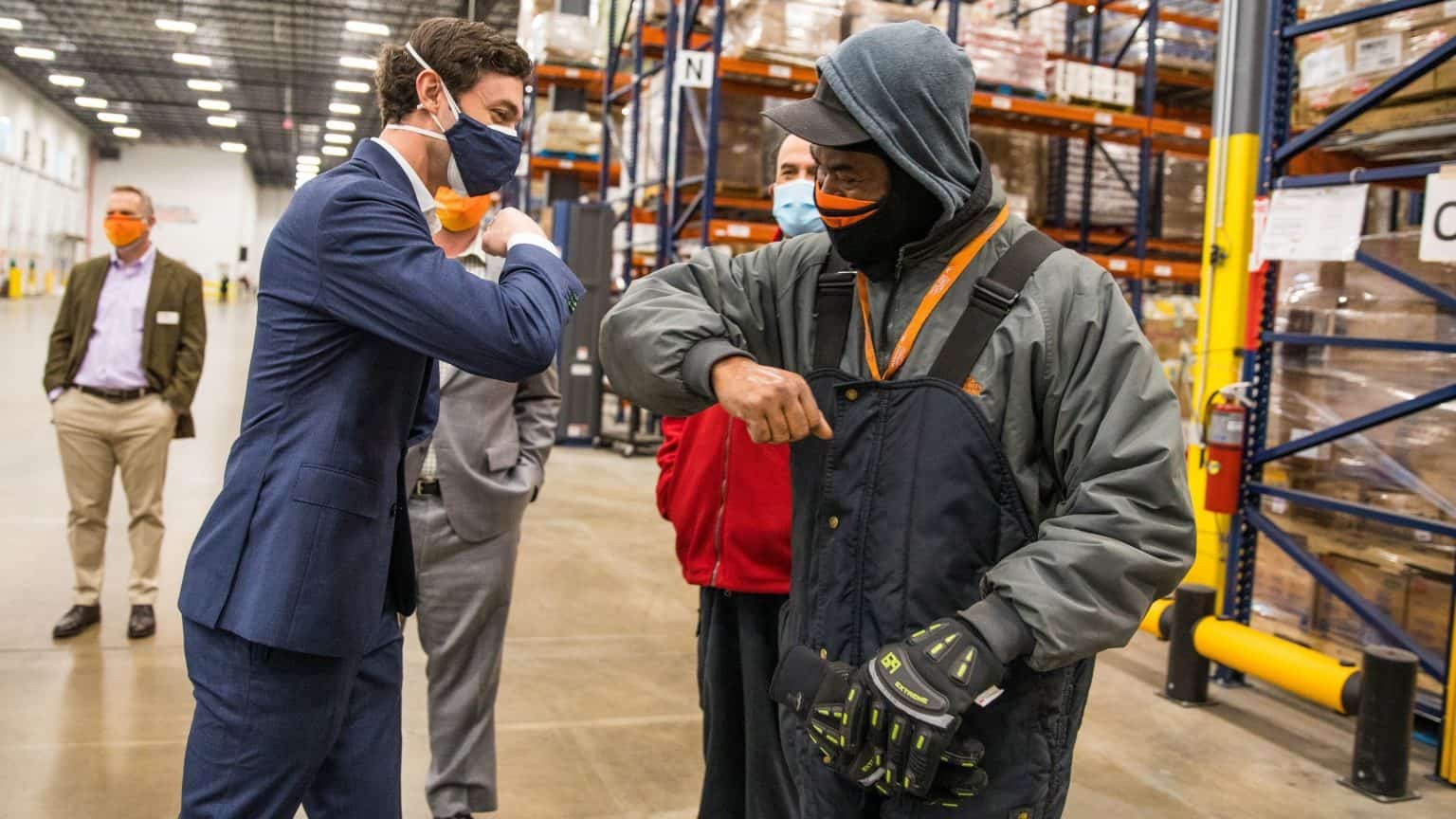 Senator Ossoff bumping elbows with a worker in a large warehouse with other staff members in the background.