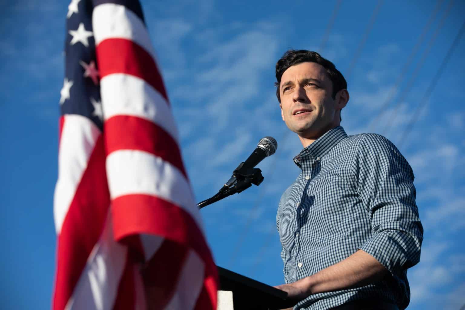 Senator Ossoff in a casual shirt under a blue sky speaking at a podium with an American flag in the foreground.